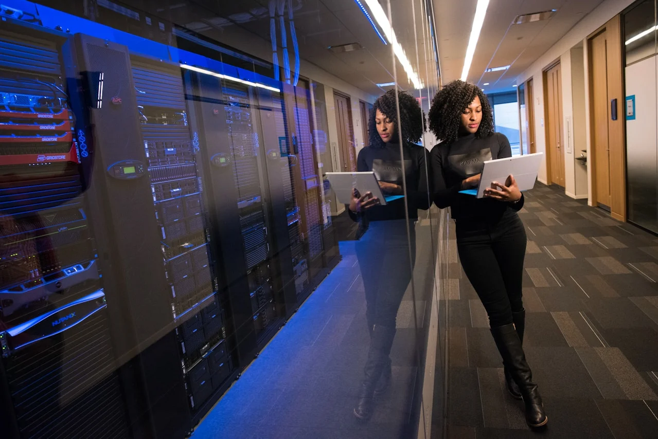 A person in a black outfit stands next to a mirrored wall, holding a laptop, with server racks illuminated in blue behind them