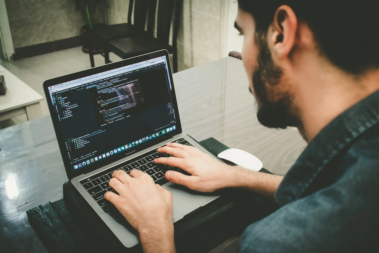 A person typing on a laptop displaying code, with a mouse beside it, in a cozy workspace with wooden furniture